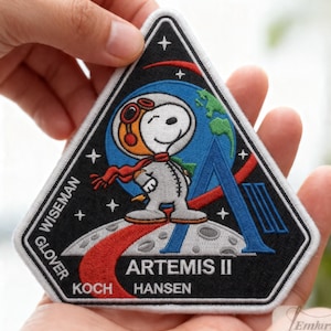 May include: Embroidered patch featuring Snoopy in an astronaut suit, part of the Artemis II mission. The patch is triangular, with a black background and white border. The design includes a blue planet, the letter "A", and the names of the crew.