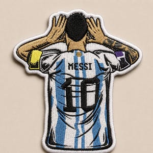 May include: An embroidered patch depicting a soccer player from behind, wearing a white and light blue striped jersey with the name MESSI and the number 10 in black. The player has hands raised to their head, with a yellow armband on the right arm.