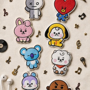 Peut inclure: Un ensemble de huit écussons brodés représentant divers personnages colorés, dont un cœur rouge, un lapin rose, un koala bleu et une créature brune, entourés de petites breloques métalliques sur le thème de la musique et d'écouteurs filaires blancs.
