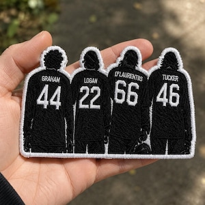 May include: An embroidered patch in black and white, depicting four figures in football jerseys. The jerseys display names and numbers: Graham 44, Logan 22, D'Laurentiis 66, and Tucker 46. The patch is held in a hand.