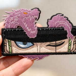May include: Embroidered patch featuring a purple dragon and a character's eyes with a black hat. The patch has a black border and is held by a hand. The eyes are blue and the character's skin is tan.