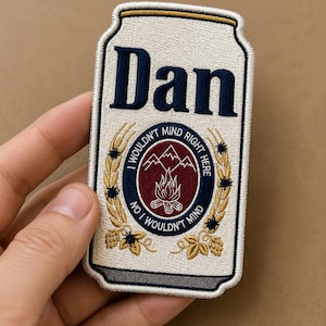 Könnte beinhalten: Gestickter Aufnäher in Form einer Bierdose mit dem Namen "Dan" in Marineblau. Der Aufnäher zeigt ein rundes Emblem mit Bergen, einem Lagerfeuer und dem Text "I WOULDN'T MIND RIGHT HERE" und "NO I WOULDN'T MIND".