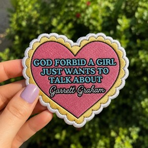May include: A heart-shaped patch with a pink centre and a scalloped yellow border. The patch features the text "GOD FORBID A GIRL JUST WANTS TO TALK ABOUT Garrett Graham" in blue and black lettering. The patch is held by a hand with light purple nail polish.
