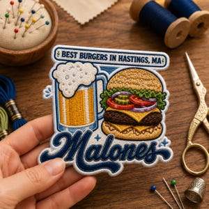 May include: Embroidered patch with a burger and beer design. The patch reads "Best Burgers in Hastings, MA" and "Malones". The burger has cheese, red onion, and pickles. The beer mug is filled with a golden liquid and topped with foam.