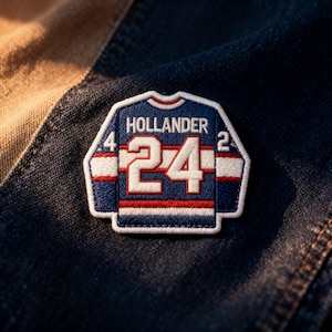 May include: An embroidered patch in the style of a hockey jersey, featuring the name "HOLLANDER" above the number "24". The patch is blue, white, and red. The number "4" is on the left and "2" on the right.