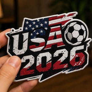 Puede incluir: Parche bordado con el texto "USA 2026" en blanco, rojo y azul, y un balón de fútbol. El diseño incorpora la bandera estadounidense, ideal para eventos deportivos o merchandising.