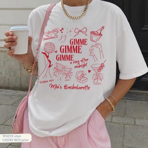 Könnte beinhalten: Weißes T-Shirt mit rotem Grafikdruck. Das Design enthält den Text "GIMME GIMME GIMME a ring after midnight" und "Mia's Bachelorette" mit Illustrationen von Cocktails, Champagner und einer Ringbox. Das Shirt hat auch den Text "Charleston, SC" und "October 2025".