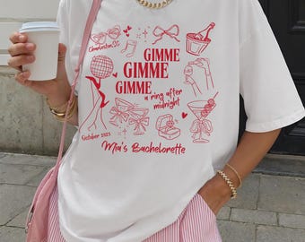 Könnte beinhalten: Weißes T-Shirt mit rotem Grafikdruck. Das Design enthält den Text "GIMME GIMME GIMME a ring after midnight" und "Mia's Bachelorette" mit Illustrationen von Cocktails, Champagner und einer Ringbox. Das Shirt hat auch den Text "Charleston, SC" und "October 2025".
