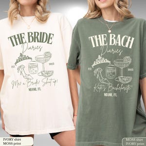 May include: Ivory and moss green t-shirts with white graphic designs. The ivory shirt says "THE BRIDE Diaries" with a tiara, shoe, cocktail, and text "Me? a Bride? Shut up!" The moss shirt says "THE BACH Diaries" with similar graphics and "Kate's Bachelorette".