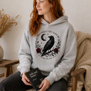 May include: A light gray hooded sweatshirt featuring a black graphic of a raven perched on a branch, surrounded by a wreath of red roses, a crescent moon, and stars, with the text "WITHOUT THE DARK WE NEVER SEE THE STARS" printed in a circle.