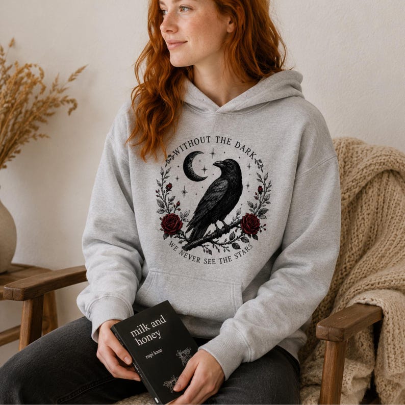May include: A light gray hooded sweatshirt featuring a black graphic of a raven perched on a branch, surrounded by a wreath of red roses, a crescent moon, and stars, with the text "WITHOUT THE DARK WE NEVER SEE THE STARS" printed in a circle.