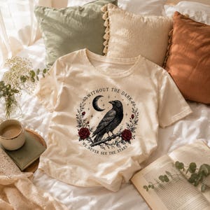 May include: A cream-colored short-sleeve t-shirt featuring a black graphic of a raven perched on a branch, surrounded by a wreath of red roses, stars, and a crescent moon. The text reads, "WITHOUT THE DARK WE NEVER SEE THE STARS."