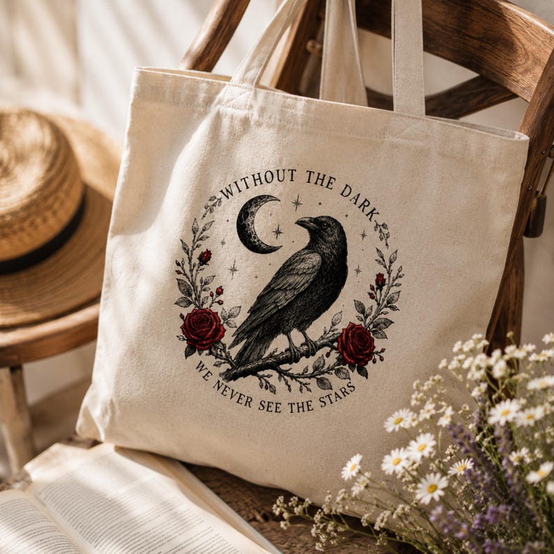 May include: A cream-colored canvas tote bag featuring a black ink illustration of a raven perched on a branch, surrounded by a wreath of red roses, a crescent moon, and stars, with the text WITHOUT THE DARK WE NEVER SEE THE STARS.