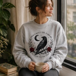 May include: A light gray crewneck sweatshirt featuring a graphic print of a black raven perched on a branch, surrounded by a circular wreath of red roses, a crescent moon, and stars, with the text "WITHOUT THE DARK WE NEVER SEE THE STARS."