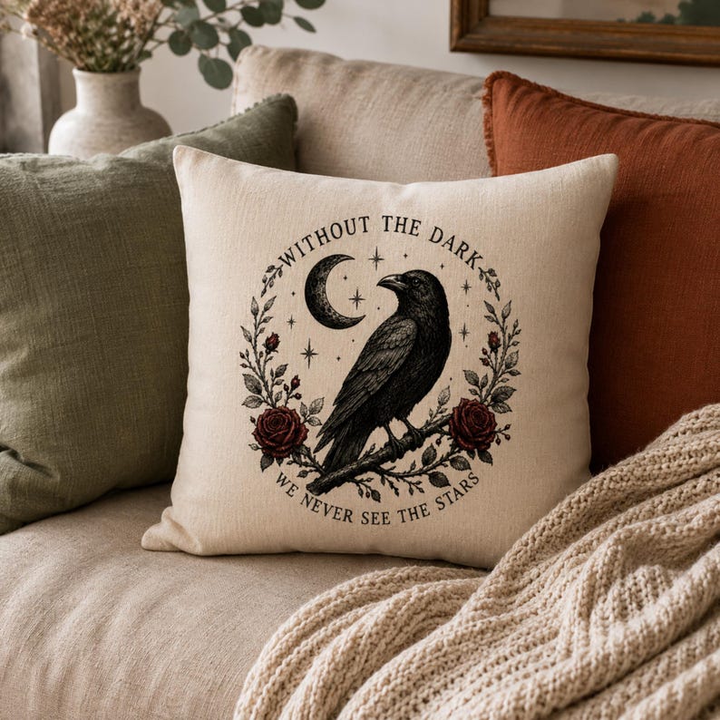 May include: A square off-white throw pillow featuring a black ink-style illustration of a raven perched on a branch, surrounded by a wreath of red roses, leaves, and a crescent moon. The text reads: WITHOUT THE DARK WE NEVER SEE THE STARS.
