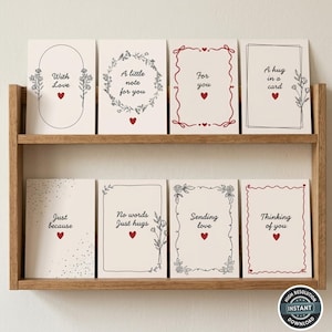 Pode incluir: Uma prateleira de madeira exibe oito cartões de felicitações com várias mensagens e designs. Cada cartão apresenta um fundo branco, bordas decorativas pretas ou vermelhas e um pequeno coração vermelho. Os cartões incluem frases como "Com amor" e "Pensando em você".