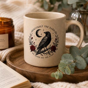 May include: A speckled beige ceramic mug featuring a black raven perched on a branch, surrounded by red roses, a crescent moon, and stars. The text "WITHOUT THE DARK WE NEVER SEE THE STARS" is printed in a circular arc around the central illustration.