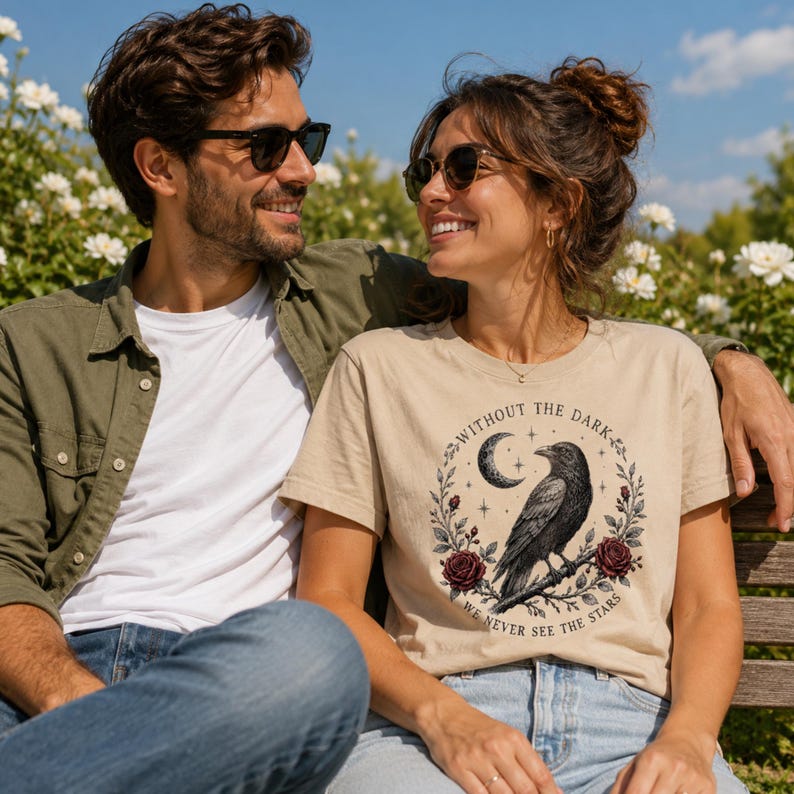 May include: A beige short-sleeved t-shirt featuring a graphic print of a black raven perched on a branch, surrounded by a crescent moon, stars, and red roses. The text "WITHOUT THE DARK, WE NEVER SEE THE STARS" is printed in a circular arc.