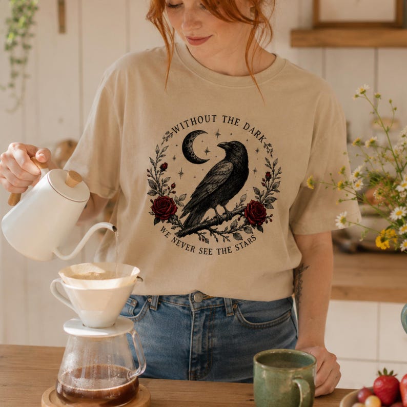 May include: A beige short-sleeve t-shirt featuring a black graphic of a raven perched on a branch, surrounded by a crescent moon, stars, and red roses. The text reads, "WITHOUT THE DARK, WE NEVER SEE THE STARS," arranged in a circular pattern.