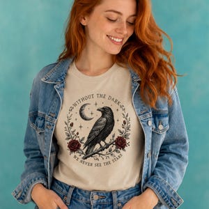 May include: A beige t-shirt featuring a black raven perched on a branch, surrounded by a circular wreath of leaves, dark red roses, a crescent moon, and stars. The text "WITHOUT THE DARK WE NEVER SEE THE STARS" is printed in a circular arc.