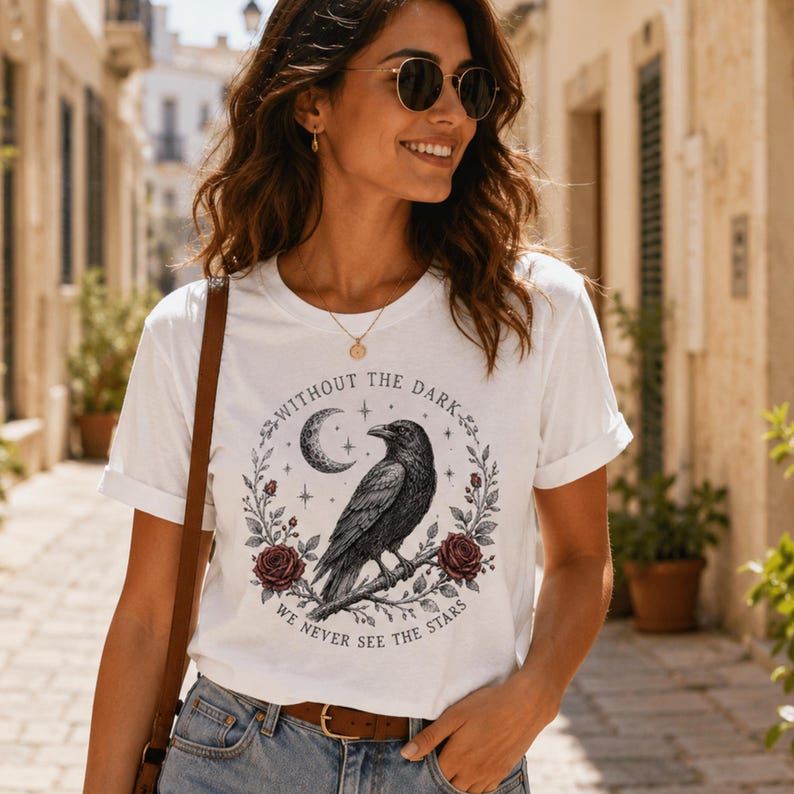 May include: A white short-sleeve t-shirt featuring a graphic print of a black raven perched on a branch, surrounded by a crescent moon, stars, and two dark red roses. The text "WITHOUT THE DARK WE NEVER SEE THE STARS" circles the illustration.