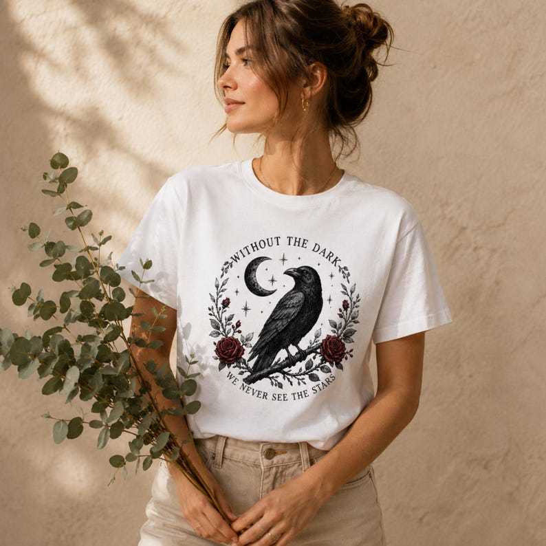 May include: A white short-sleeved t-shirt featuring a black graphic of a raven perched on a branch, surrounded by a circular wreath of dark red roses, a crescent moon, and stars, with the text "WITHOUT THE DARK WE NEVER SEE THE STARS".