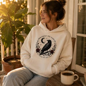 May include: A cream-colored hooded sweatshirt featuring a graphic print of a black raven perched on a branch, surrounded by red roses and a crescent moon. The text reads, "WITHOUT THE DARK WE NEVER SEE THE STARS" in a circular arrangement.