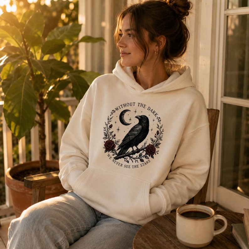 May include: A cream-colored hooded sweatshirt featuring a graphic print of a black raven perched on a branch, surrounded by red roses and a crescent moon. The text reads, "WITHOUT THE DARK WE NEVER SEE THE STARS" in a circular arrangement.
