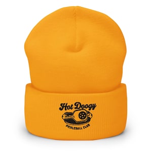 May include: Mustard yellow beanie with a folded brim. The front of the brim features the text "Hot Doogy Pickleball Club" in black, along with a graphic of a pickleball paddle and ball.