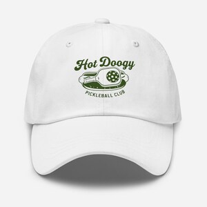 May include: White baseball cap with a curved brim. The cap features a green graphic of a pickleball paddle and ball, with the words "Hot Doogy Pickleball Club" in green lettering. The cap is a classic, adjustable style.