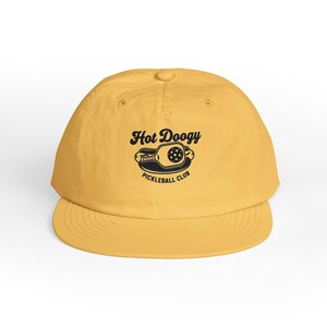 May include: Mustard yellow baseball cap with a curved brim. The cap features the text "Hot Doogy Pickleball Club" in black, along with a graphic of a pickleball paddle and ball. The cap is made of a soft material and has a rounded crown.