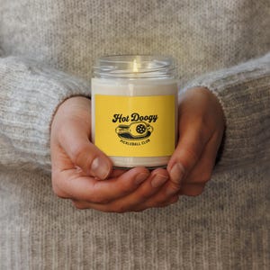 May include: A lit candle in a clear glass jar with a yellow label. The label features the text "Hot Doogy Pickleball Club" in black and white. The jar is held in both hands.
