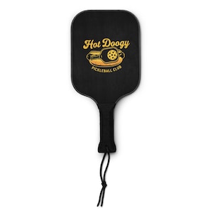 May include: Black pickleball paddle with a yellow graphic that reads "Hot Doogy Pickleball Club." The paddle has a black handle with a black cord attached. The paddle is designed for playing pickleball.
