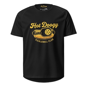 May include: Black t-shirt with a yellow graphic that reads "Hot Doogy Pickleball Club." The graphic features a pickleball paddle and ball inside a hot dog bun. The shirt has a crew neck and short sleeves.