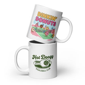 May include: Two white ceramic mugs stacked on top of each other. The top mug features the text "Dinkin' Donuts" with cartoon donuts playing pickleball. The bottom mug has the text "Hot Doogy Pickleball Club" with a pickleball paddle graphic.