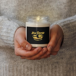 May include: A lit candle in a clear glass jar with a black label. The label features the text "Hot Doogy Pickleball Club" in gold lettering and a pickleball paddle graphic. The candle is held in both hands.