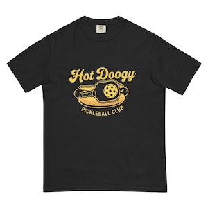 May include: Black t-shirt with a yellow graphic that reads "Hot Doogy Pickleball Club." The graphic features a pickleball paddle and ball inside a hot dog bun. The shirt is a comfortable, casual style.