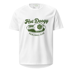 May include: White athletic t-shirt with a green graphic that reads "Hot Doogy Pickleball Club." The graphic features a pickleball paddle and ball. The shirt has short sleeves and a crew neck.
