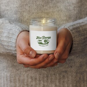 May include: A lit candle in a clear glass jar with a white label. The label features the text "Hot Doogy Pickleball Club" in green. The candle is held in both hands.