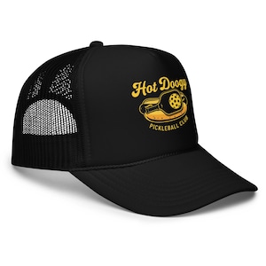May include: Black trucker hat with a mesh back and a curved brim. The hat features the text "Hot Doogy Pickleball Club" in yellow, along with a graphic of a pickleball paddle and ball.