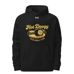 May include: Black hooded sweatshirt with a front pocket. The sweatshirt features a yellow and black graphic of a pickleball paddle and ball with the text "Hot Doogy Pickleball Club". The Under Armour logo is visible.