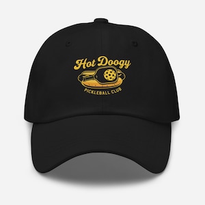 May include: Black baseball cap with a yellow graphic that reads "Hot Doogy Pickleball Club." The graphic features a pickleball paddle and ball inside a hot dog bun. The hat has a curved brim and a button on top.