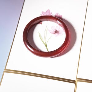May include: A dark red bangle bracelet. The bracelet is circular and sits on a white surface with a floral design. The floral design features pink flowers and green leaves. The background has a gradient of purple and white.