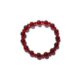 May include: A red beaded bracelet with a mix of large and small round beads. The bracelet is made of a shiny, translucent material and is displayed on a white background. The beads are a deep, rich red color.
