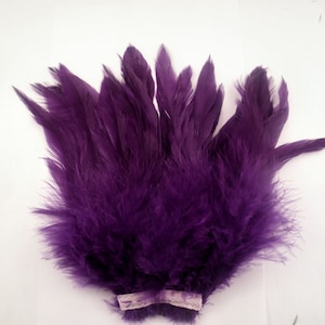 May include: A cluster of deep purple feathers, likely for crafting or costume design. The feathers are a rich, saturated color, with a soft, fluffy texture at the base and more defined tips. A light-colored fabric strip secures the base of the feathers.