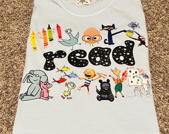 Puede incluir: Camiseta azul claro con la palabra "read" en letras negras con estrellas blancas. La camiseta presenta ilustraciones de dibujos animados coloridos de crayones, animales y personajes de libros infantiles.