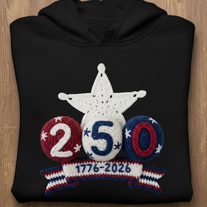Puede incluir: Sudadera con capucha negra con una estrella blanca y los números "250" en un diseño de ganchillo. Los números son en rojo, blanco y azul, con el texto "1776-2026" en una pancarta debajo. La sudadera está hecha de un material suave.