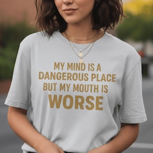 Puede incluir: Camiseta gris claro con el texto dorado: "MY MIND IS A DANGEROUS PLACE BUT MY MOUTH IS WORSE". La camiseta, de cuello redondo, parece estar hecha de un material suave. El texto está en negrita.