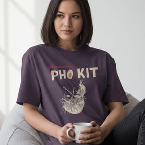 Könnte beinhalten: Ein dunkelviolettes T-Shirt mit den Worten "PHO KIT" in großen, weißen Großbuchstaben. Darunter befindet sich eine Grafik einer Schüssel Pho mit Essstäbchen. Die Person hält einen weißen Becher.
