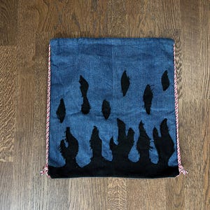Peut inclure: Un sac en jean bleu avec un motif de flammes noires. Le sac a un cordon de serrage en corde rouge et blanche et un motif de flammes noires sur le devant.